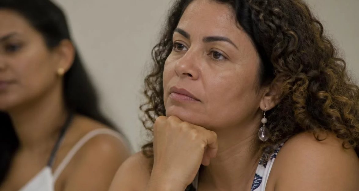 Participante com o braço no alto pedindo a vez, diante de folhas rabiscadas espalhadas pela mesa